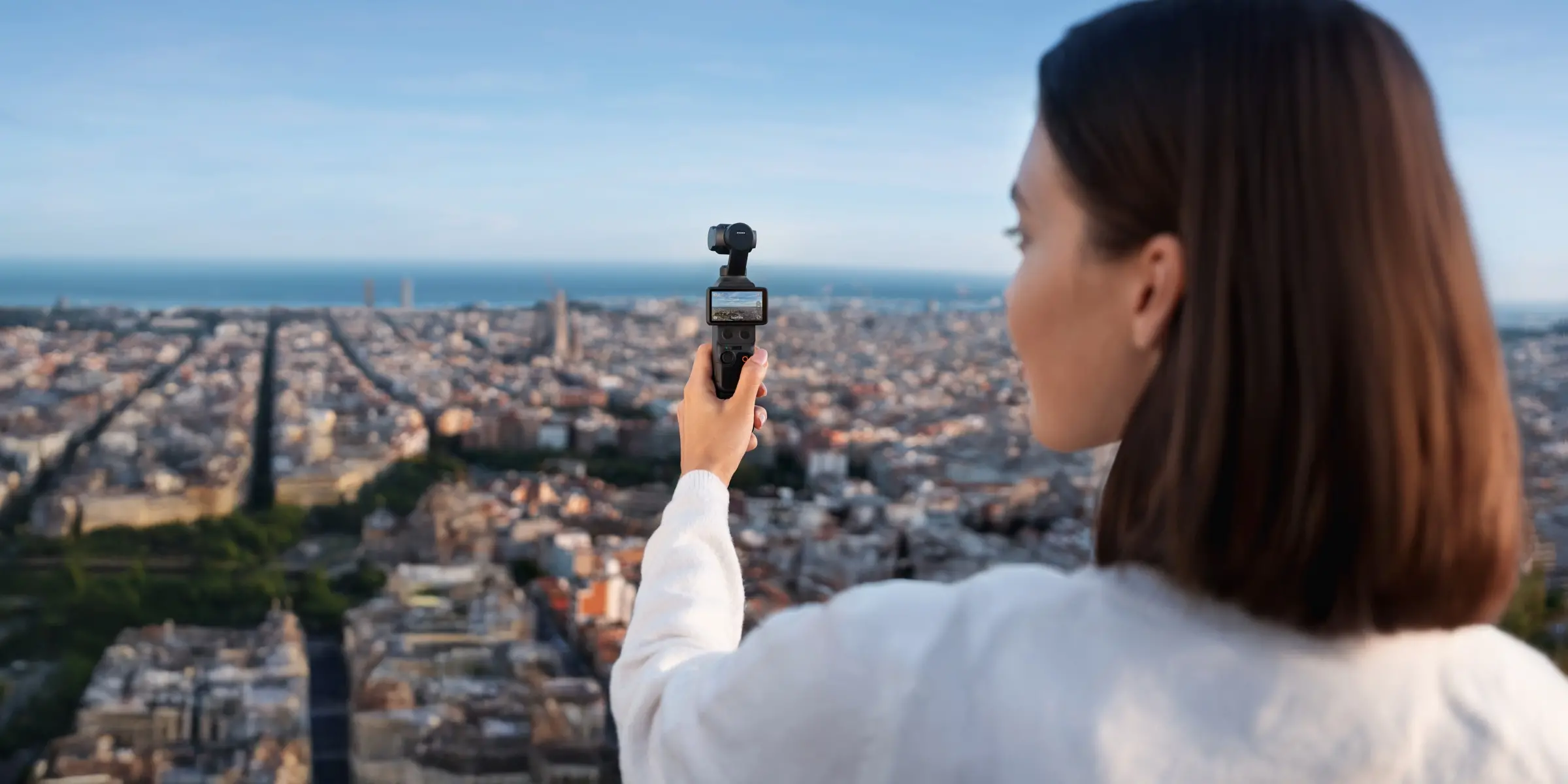 A person holding the DJI Osmo Pocket 4 gimbal camera at arm's length above a city skyline, with the camera's small vertical screen and three-axis gimbal head visible